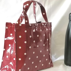 Peut inclure: Un sac fourre-tout bordeaux brillant &agrave; pois blancs. Le sac a deux poign&eacute;es et une forme rectangulaire. Une bouteille noire est visible en arri&egrave;re-plan, sur fond blanc.