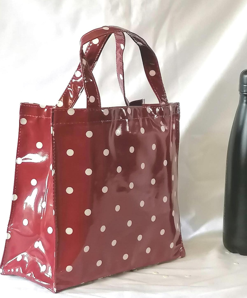 Peut inclure: Un sac fourre-tout bordeaux brillant &agrave; pois blancs. Le sac a deux poign&eacute;es et une forme rectangulaire. Une bouteille noire est visible en arri&egrave;re-plan, sur fond blanc.
