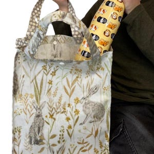 Peut inclure: Un sac fourre-tout avec un fond bleu clair pr&eacute;sentant des illustrations de li&egrave;vres et de feuillage. Le sac a une poign&eacute;e &agrave; pois et contient une gourde jaune avec des illustrations de cochons d'Inde. Le sac mesure environ 30 cm de large.