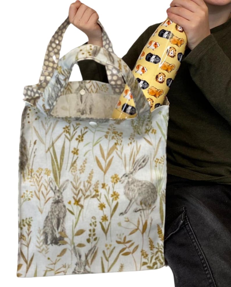 Peut inclure: Un sac fourre-tout avec un fond bleu clair pr&eacute;sentant des illustrations de li&egrave;vres et de feuillage. Le sac a une poign&eacute;e &agrave; pois et contient une gourde jaune avec des illustrations de cochons d'Inde. Le sac mesure environ 30 cm de large.