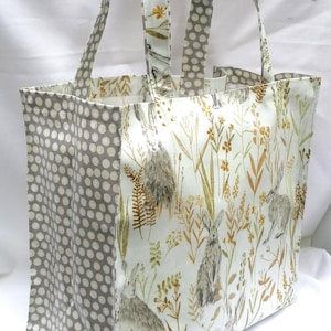 Op de afbeelding: Een rechthoekige draagtas met een ontwerp met een natuurthema. De tas heeft een witte achtergrond met illustraties van hazen, planten en bloemen in bruin- en groentinten. De zijkanten zijn grijs met witte stippen.