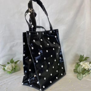 Peut inclure: Sac fourre-tout noir &agrave; pois blancs. Le sac a deux poign&eacute;es et une finition brillante. Le sac est rectangulaire avec un fond plat et convient au transport de divers articles.