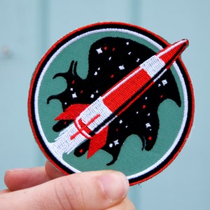 Può includere: Una toppa rotonda ricamata di colore verde con un bordo bianco e un razzo rosso e bianco che vola attraverso una sagoma nera di una creatura con stelle.