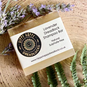 Könnte beinhalten: Ein rechteckiger Lavendel-Dreadlock-Shampoo-Riegel mit weißem Etikett und einem schwarz-goldenen Logo. Der Riegel ist natürlich und sulfatfrei. Lavendelzweige und grüne Farnblätter umgeben den Riegel auf einer Holzoberfläche.