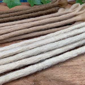 Puede incluir: Primer plano de un paquete de rastas sintéticas en tres tonos de marrón, beige y blanco. Las rastas están hechas de un material suave y texturizado y están listas para ser añadidas a tu cabello.