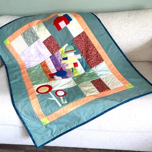 Peut inclure: Une courtepointe faite à la main avec un motif abstrait. Elle présente des carrés de tissu aux couleurs vives : bleu, vert, orange, rouge et violet. Bordée de tissu turquoise et d'une bordure bleu foncé, elle est posée sur un canapé blanc.