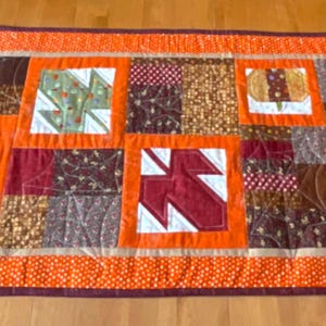 Puede incluir: Un camino de mesa acolchado rectangular con un borde naranja y un diseño de patchwork. El patchwork presenta cuadrados con motivos de temática otoñal, incluyendo calabazas y hojas, en tonos naranja, marrón y verde. Mide aproximadamente 30 cm de ancho y 91 cm de largo.