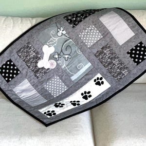 May include: A grey and white pet blanket with a black border. The blanket features a bone and paw print design, with various patterns including polka dots and stripes. The blanket is made of quilted fabric.