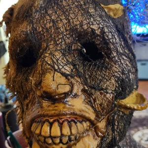 May include: A creepy scarecrow mask with a burlap and mesh covering, a wide, toothy grin, and a pair of small, pointed ears.