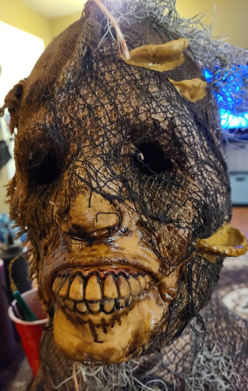 May include: A creepy scarecrow mask with a burlap and mesh covering, a wide, toothy grin, and a pair of small, pointed ears.