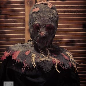 May include: A creepy scarecrow mask with a tattered burlap face covering and a red and black burlap collar. The mask is made of a realistic material and has a distressed look.