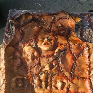 May include: A brown leather vest with multiple stitched-on human faces. The faces are all different shapes and sizes, and some have their mouths open. The vest is made of a patchwork of different pieces of leather, and the stitching is visible.
