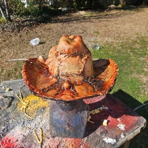 May include: A brown leather cowboy hat with a stitched design and blood-like stains. The hat is on a display stand.