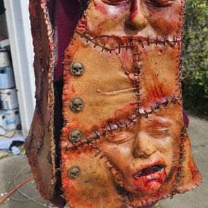 May include: A brown leather vest with stitched seams and a bloody, grotesque human face sewn onto the front. The vest has several skull-shaped buttons.