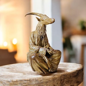 Puede incluir: Una escultura de color bronce de una figura con cabeza de cabra en posición sentada, vestida con una túnica drapeada. La cabra tiene cuernos curvados y una larga barba. La escultura está colocada sobre una superficie de madera rústica.