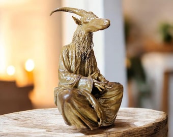 May include: A bronze-colored sculpture of a goat-headed figure in a seated position, wearing a draped robe. The goat has curved horns and a long beard. The sculpture is placed on a rustic wooden surface.