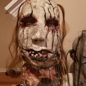 May include: A white and gray mask with black lines and red paint. The mask has a realistic human face with a wide mouth and teeth. The mask is attached to a brown base with long brown hair.