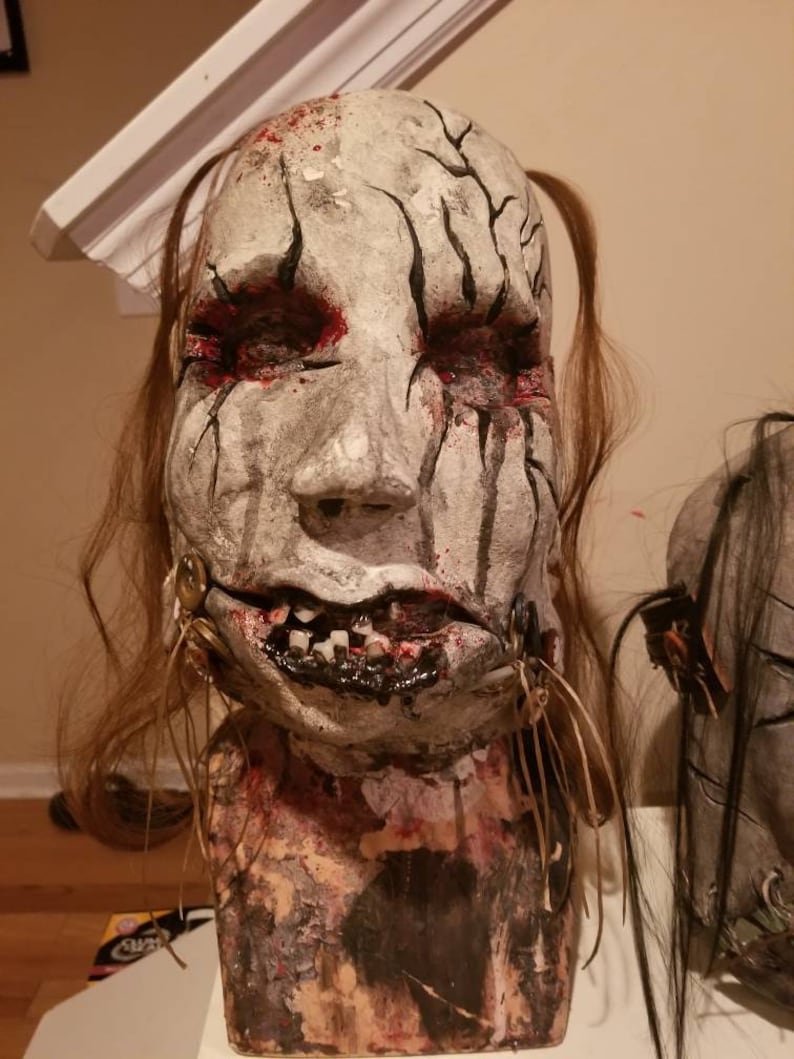 May include: A white and gray mask with black lines and red paint. The mask has a realistic human face with a wide mouth and teeth. The mask is attached to a brown base with long brown hair.