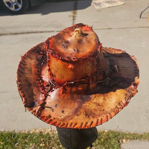 May include: A brown leather cowboy hat with orange stitching and a distressed look. The hat is on a stand and has a unique design.