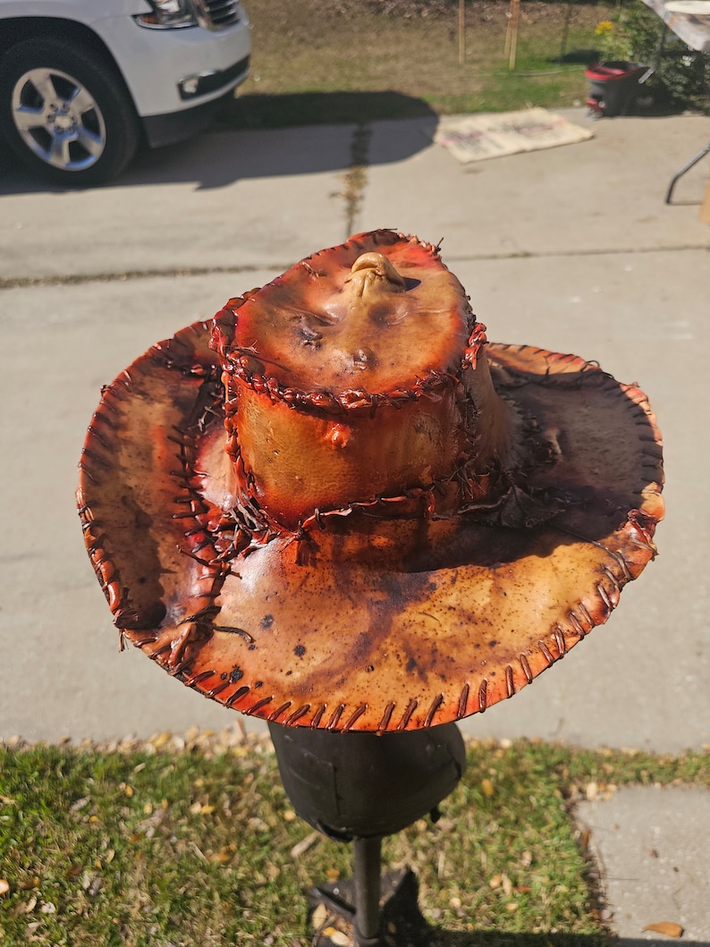May include: A brown leather cowboy hat with orange stitching and a distressed look. The hat is on a stand and has a unique design.