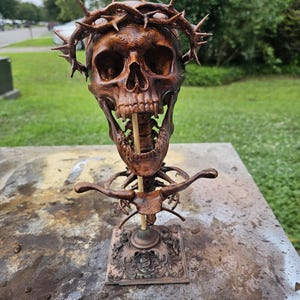 Puede incluir: Una escultura de cráneo marrón detallada con una corona de espinas sobre el cráneo. El cráneo está montado en una varilla vertical unida a una base cuadrada ornamentada con intrincados relieves, mostrando un acabado oscuro y desgastado.