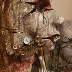 May include: A creepy, realistic-looking, handmade doll head with a cracked, gray face, a bloody eye, and a stitched-up mouth. The doll's hair is long and brown, and it has several buttons sewn onto its face and neck.