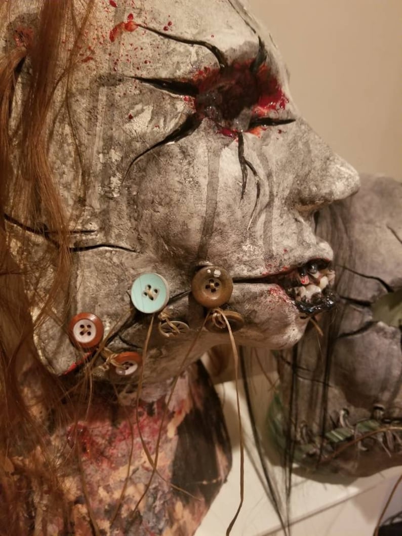 May include: A creepy, realistic-looking, handmade doll head with a cracked, gray face, a bloody eye, and a stitched-up mouth. The doll's hair is long and brown, and it has several buttons sewn onto its face and neck.