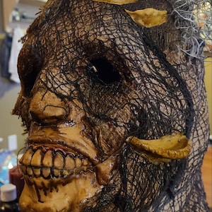 May include: A creepy, detailed Halloween mask with a burlap-like texture and a stitched-on mouth. The mask has a dark, weathered look and features a single eye hole.