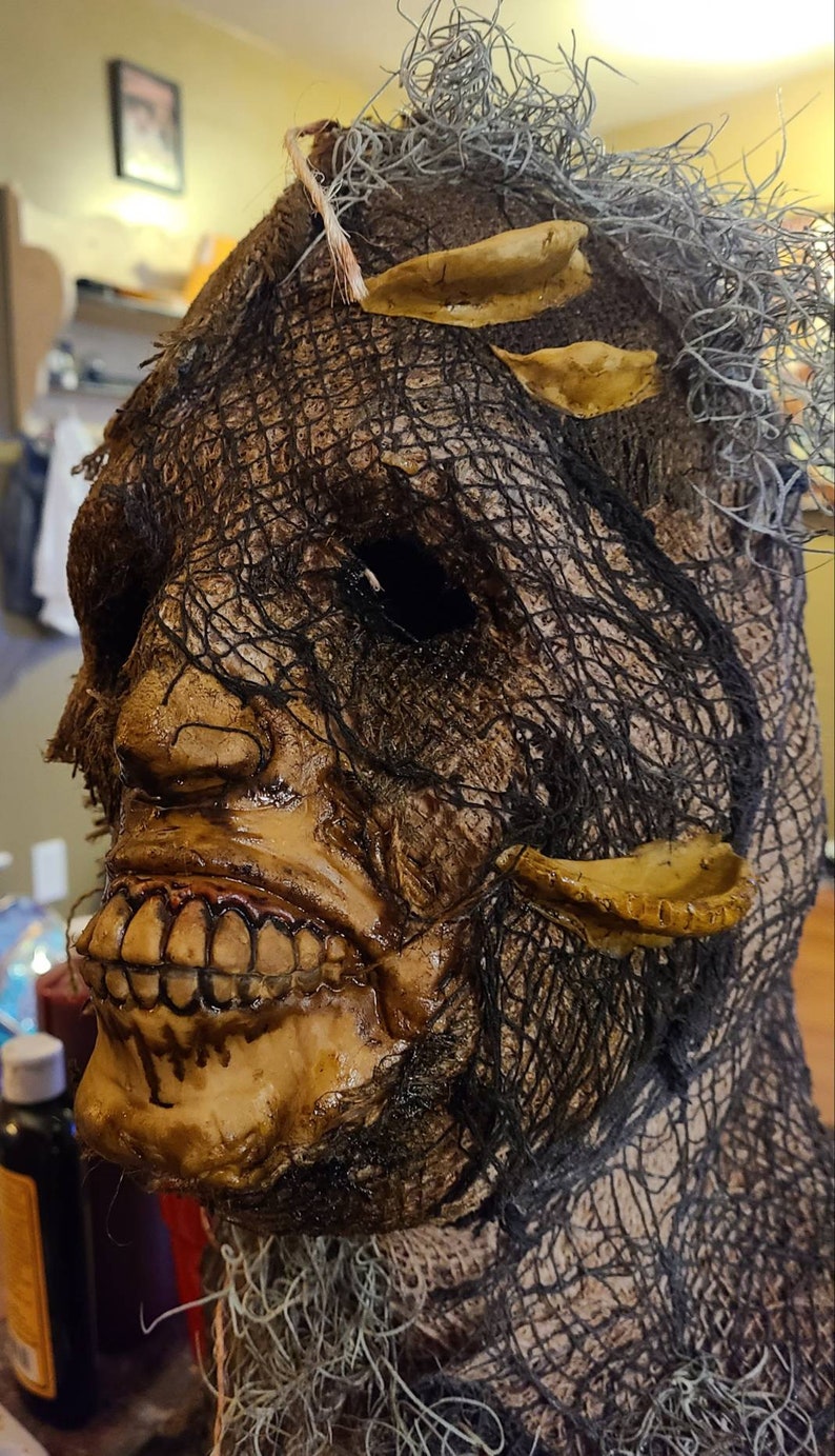 May include: A creepy, detailed Halloween mask with a burlap-like texture and a stitched-on mouth. The mask has a dark, weathered look and features a single eye hole.