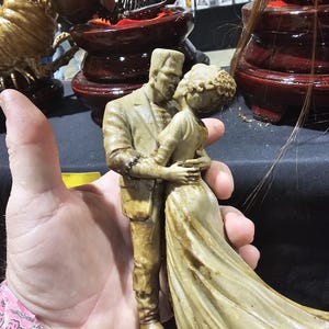 K&ouml;nnte beinhalten: Beige Figur von Frankenstein und seiner Braut, die sich umarmen. Die Figuren sind detailliert, mit dem flie&szlig;enden Kleid der Braut und den ikonischen Merkmalen von Frankenstein. Die Figur wird in einer Hand gehalten, mit anderen dekorativen Gegenst&auml;nden im Hintergrund.