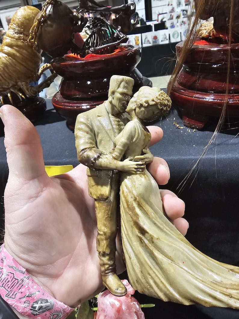 K&ouml;nnte beinhalten: Beige Figur von Frankenstein und seiner Braut, die sich umarmen. Die Figuren sind detailliert, mit dem flie&szlig;enden Kleid der Braut und den ikonischen Merkmalen von Frankenstein. Die Figur wird in einer Hand gehalten, mit anderen dekorativen Gegenst&auml;nden im Hintergrund.