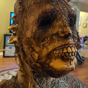 May include: A creepy, realistic-looking Halloween mask made of burlap and mesh. The mask has a grotesque, decaying face with a wide, toothy grin. The mask is perfect for adding a touch of horror to any Halloween costume.