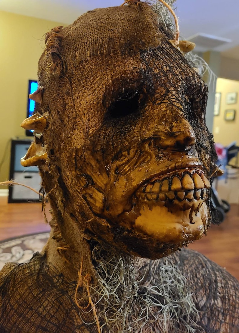 May include: A creepy, realistic-looking Halloween mask made of burlap and mesh. The mask has a grotesque, decaying face with a wide, toothy grin. The mask is perfect for adding a touch of horror to any Halloween costume.