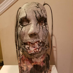 May include: A realistic, detailed, gray and black horror mask with stitched lips and exposed teeth. The mask has long black hair and is mounted on a textured, brown base.
