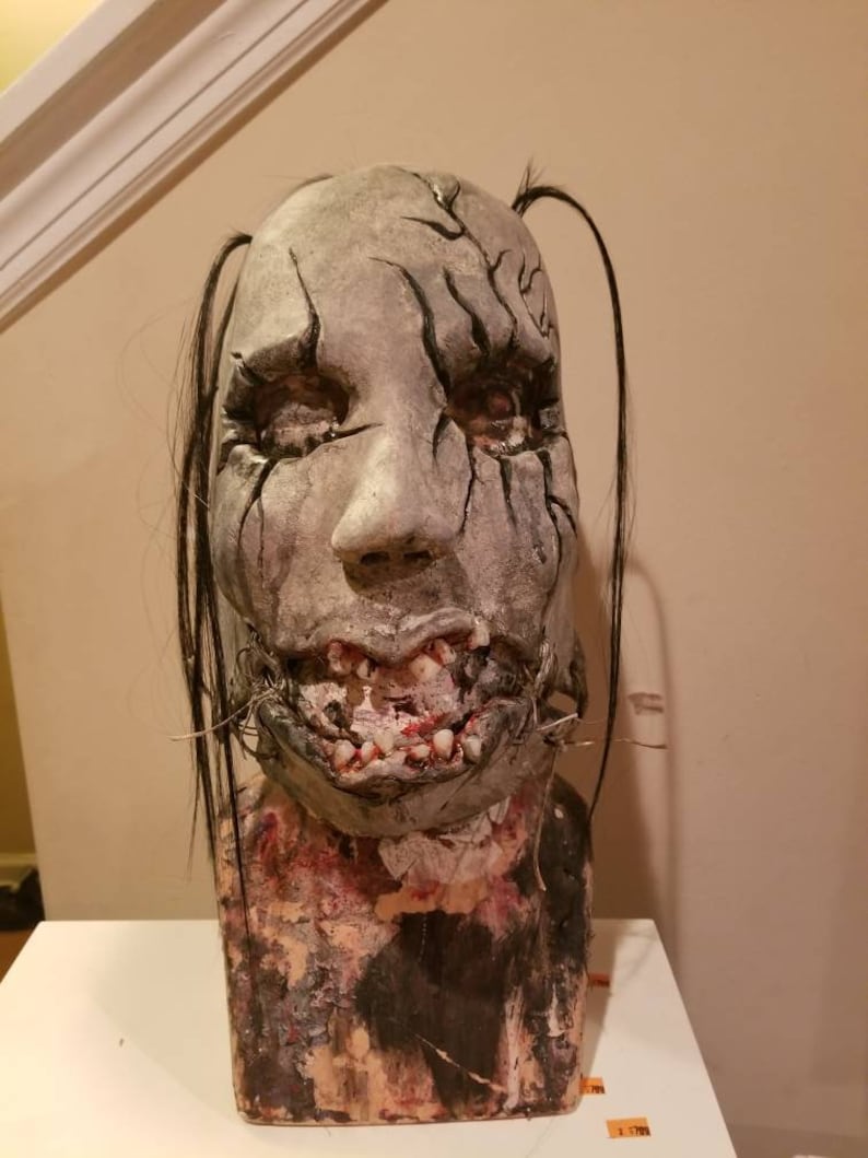 May include: A realistic, detailed, gray and black horror mask with stitched lips and exposed teeth. The mask has long black hair and is mounted on a textured, brown base.