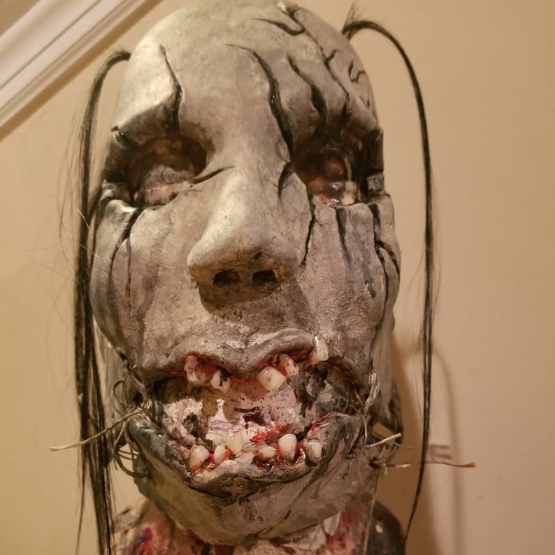 May include: A detailed, realistic horror mask with a cracked, gray face, black hair, and a wide, toothy grin. The mask has a stitched mouth and a realistic texture.