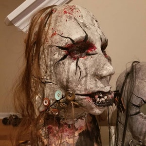 May include: A white and gray mask with a stitched mouth and buttons. The mask has red paint splatters and black lines around the eyes. The mask is on a mannequin head with long brown hair.