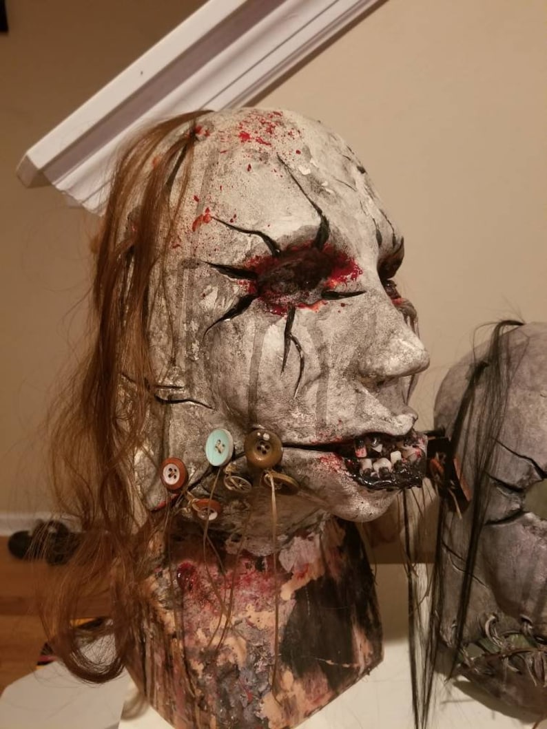 May include: A white and gray mask with a stitched mouth and buttons. The mask has red paint splatters and black lines around the eyes. The mask is on a mannequin head with long brown hair.