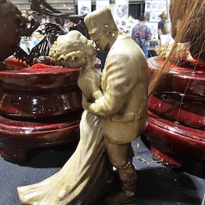 K&ouml;nnte beinhalten: Eine beigefarbene Skulptur von Frankenstein und seiner Braut, die sich umarmen und k&uuml;ssen. Die Figuren sind detailliert, die Braut in einem flie&szlig;enden Kleid und das Monster in einem Anzug. Die Skulptur steht auf einem dunkelroten, mehrstufigen Sockel.