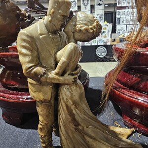 K&ouml;nnte beinhalten: Eine beigefarbene Skulptur von Frankenstein, der seine Braut umarmt. Die Figuren sind detailliert, die Braut in einem flie&szlig;enden Kleid und das Monster in einem Anzug. Die Skulptur steht auf einem dunkelroten, lackierten Sockel, mit anderen &auml;hnlichen Skulpturen im Hintergrund.