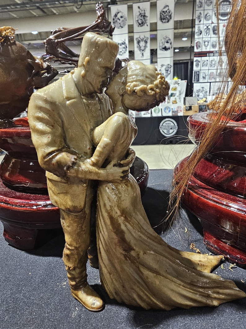 K&ouml;nnte beinhalten: Eine beigefarbene Skulptur von Frankenstein, der seine Braut umarmt. Die Figuren sind detailliert, die Braut in einem flie&szlig;enden Kleid und das Monster in einem Anzug. Die Skulptur steht auf einem dunkelroten, lackierten Sockel, mit anderen &auml;hnlichen Skulpturen im Hintergrund.