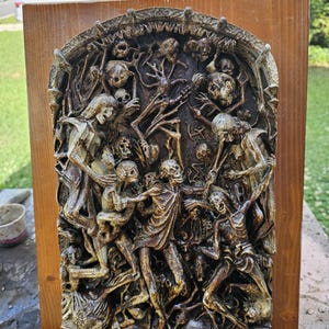 May include: A rectangular wooden plaque featuring a dark, relief-carved sculpture of multiple skeletal figures in an arched frame. The intricate, textured brown and black carving depicts a dense, chaotic scene of interconnected human skeletons.
