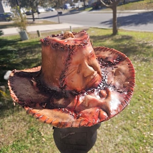 May include: A brown leather cowboy hat with a stitched design and a human face sculpted into the crown. The hat is stained with red and brown paint, giving it a bloody and distressed look.