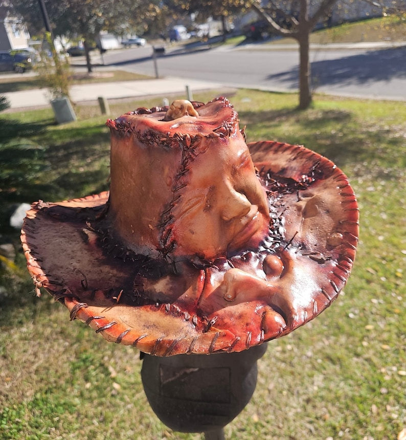 May include: A brown leather cowboy hat with a stitched design and a human face sculpted into the crown. The hat is stained with red and brown paint, giving it a bloody and distressed look.