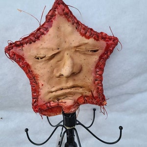 May include: A star-shaped, handmade art piece with a face, mounted on a black metal stand. The face has a neutral expression, with a stitched red border. The art piece is a unique, handcrafted item.