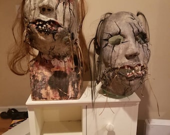 May include: Two handmade horror masks with stitched mouths and cracked paint. The masks are displayed on a white shelf unit with two drawers.