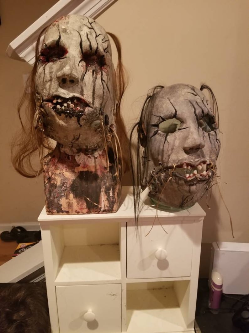 May include: Two handmade horror masks with stitched mouths and cracked paint. The masks are displayed on a white shelf unit with two drawers.