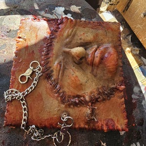 Op de afbeelding: Een vierkant, getextureerd kunstwerk in vleestint met een gestikte rand. Een zilveren ketting en sleutelringen zijn bevestigd. Het kunstwerk heeft een verhoogde, organische vorm en is bevlekt met rode tinten.