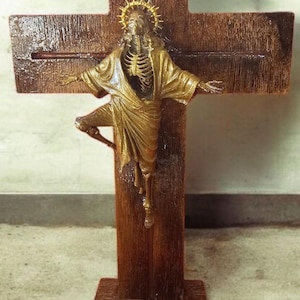 May include: A wooden crucifix with a skeletal figure in a gold robe. The figure has a gold halo and is attached to a dark brown cross. The cross is mounted on a wooden base, creating a unique religious art piece.