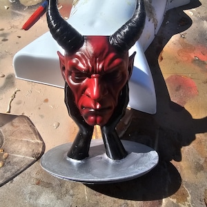 May include: A red devil head sculpture with black horns and hands supporting the chin. The sculpture is on a silver-colored base. The devil's face is detailed with a stern expression. The sculpture is placed in front of a white mannequin torso.